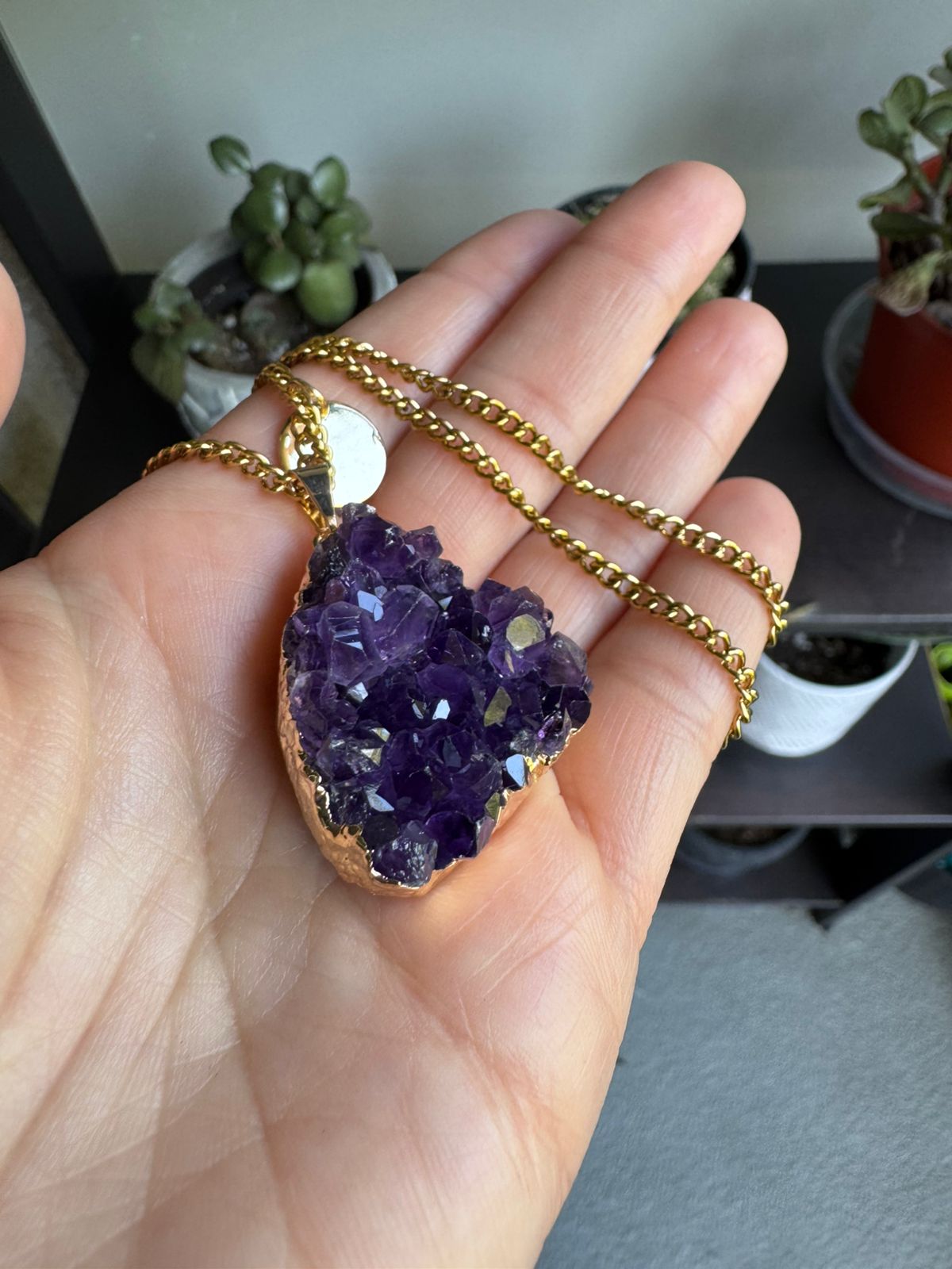 Hand holding a purple amethyst pendant on a gold chain with a blurred indoor background